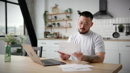 Man Working on Laptop with Documents in Kitchen