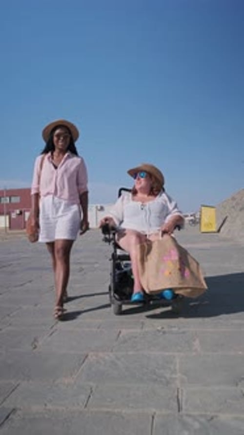 Woman Pushing Friend in Wheelchair on Beach Boardwalk in Slow Motion