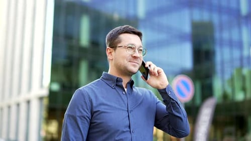 Smiling businessman talking on mobile phone while walking on street near office building. Handsome