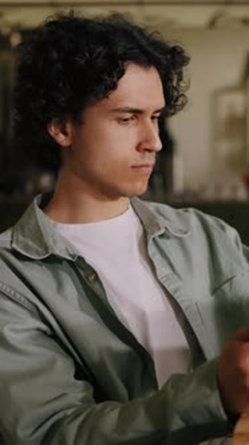 Thoughtful Young Man with Curly Hair Sitting Indoors in Casual Shirt Focusing Intently As He Looks