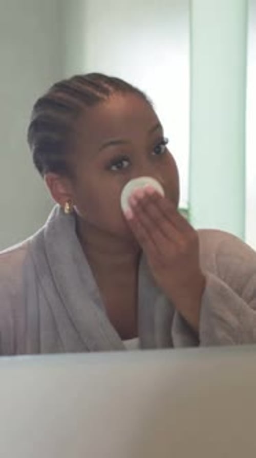 Vertical video: Glancing at mirror, woman in robe removing makeup at vanity with cotton pad