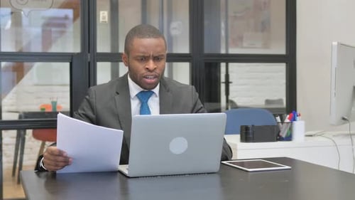 Frustrated Businessman Reading Documents and Using Laptop