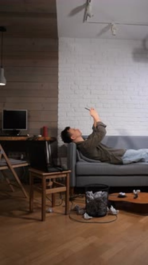 Vertical Video Man Lying on a Sofa Looking at His Smartphone Procrastinating and Experiencing a