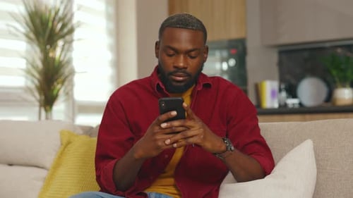 Close Up of Young African American Man Use Phone Look Around Sit on Sofa in Home Communication in
