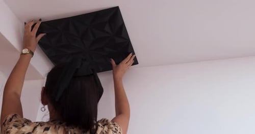 Joven morena caucásica con el pelo atado y un reloj dorado que pega un plafón negro sobre un techo blanco,