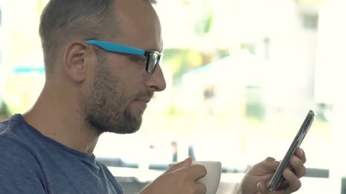 Young Man Texting on Smartphone and Drinking Coffee in Cafe 30s