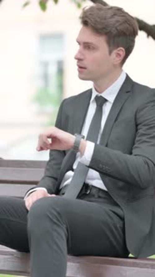 Man in Suit Sits Checking Smartwatch Outdoors