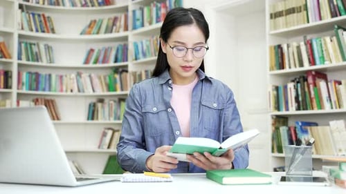Focused asian female student learning reading a book in university campus library space. Young woman