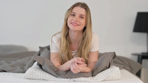 Smiling Young Woman Looking at Camera while Lying on Stomach in Bed