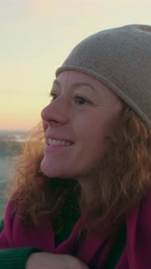 Smiling Woman with Curly Hair Wearing a Hat and Scarf Enjoying a Sunset By the Beach