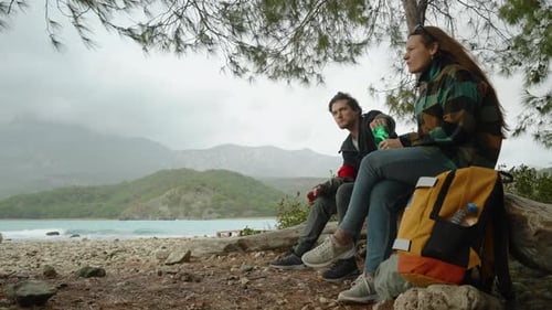 Traveling couple, a man and a woman, have stopped to have a snack while sitting on a log.