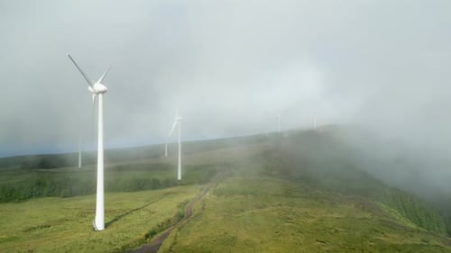 Wind turbines spin on misty morning on lush mountain ridge, aerial riser