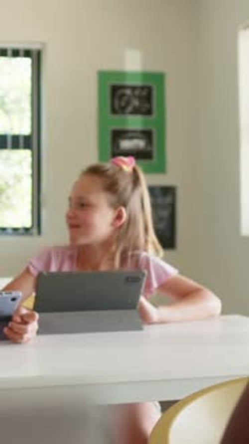 Turning toward peer entering left, seated girl with scrunchie holding tablet and phone in classroom