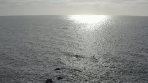 Grey ocean aerial ascends, retreats from SUP paddleboarder near rocks