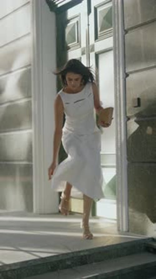 Jovem mulher de vestido branco sai do prédio em dia ensolarado