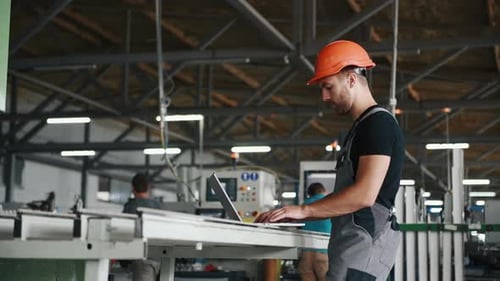 Worker in uniform stands in the factory and typing on laptop