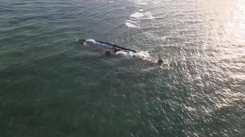 Drone aerial moving slowly over waves crashing into a shipwreck during sunset