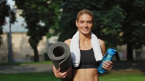 Female Holding Twisted Black Yoga Mat Bottle of Water and White Towel in Park