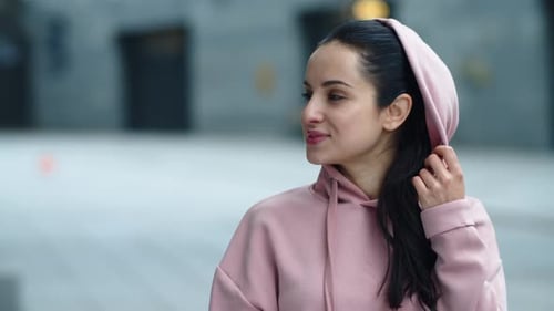 Smiling sportswoman puts on hoodie and poses on city street in slow motion