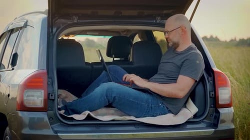 Empresário contemporâneo concentrado trabalhando remotamente digitando em um laptop sentado no porta-malas do carro