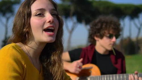 junge, sorglose Frau sitzt auf dem Rasen und singt mit ihrer Freundin, die Gitarre spielt