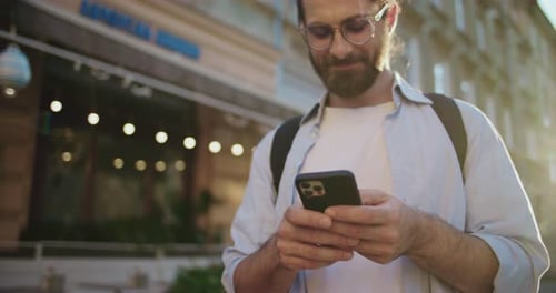 Portrait of Handsome Focused Young Bearded Man Standing in City Center Texting with Smartphone