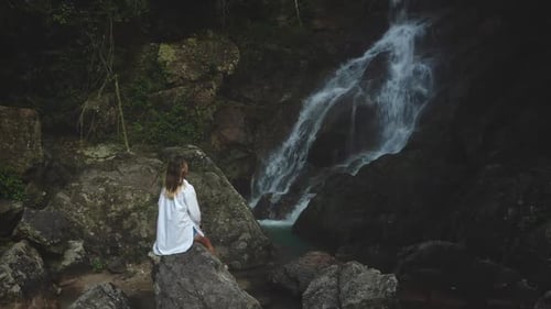 Woman Sitting on Rocks by Tropical Waterfall