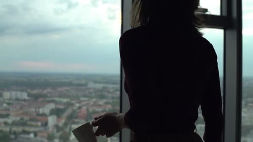 Woman Drinks Coffee Overlooking City from Highrise Apartment