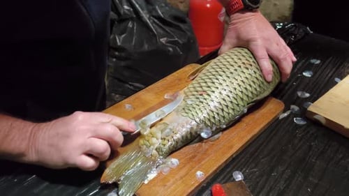 Hands scrape scales from a large fish on a wooden cutting board, removing them with firm strokes in