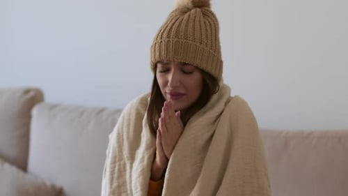 Jeune femme froide enveloppée dans une couverture avec un bonnet tricoté sur la tête, gelée à la maison, la réchauffant