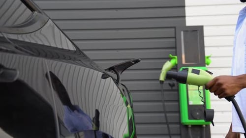 Black Man Plugging in Charging Cable to Electric Vehicle and Charges Batteries at Station