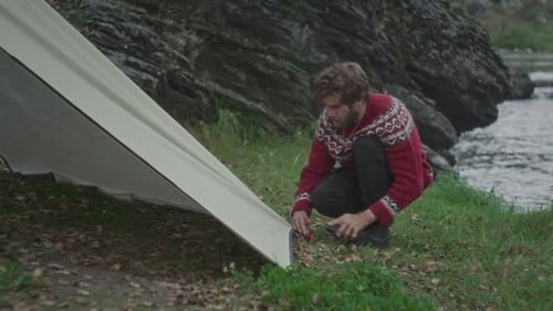Tourist Securing Tent Corner with Stone on Riverbank Campsite