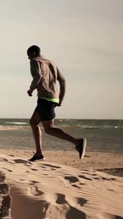 Vertical video: Sprinting man starting uphill vaulting wall and landing in sand, training in hoodie