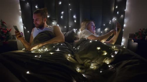 Young Couple Looks at Phones in Bed at Night