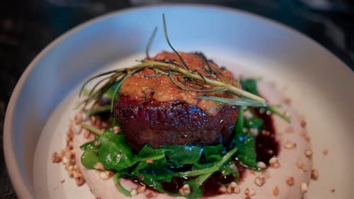 Close up of a gourmet meat dish served with sauce, greens, and rosemary garnish on a white plate