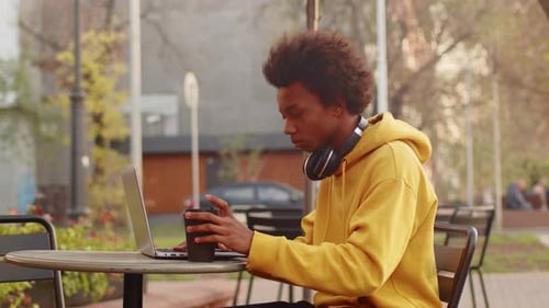 Young Black Programmer Drinking Coffee From a Paper Cup and Working on a Laptop Sitting at the Table