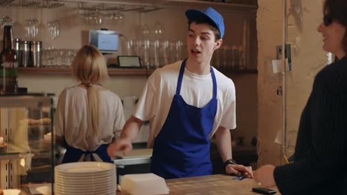 A Salesman in a Cafe Smilingly Serves a Female Customer Standing Behind the Counter and Pointing to