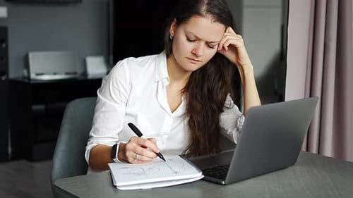 Young Tired Woman Using Laptop in Home Workplace Writing Notes Female Student Learning and Watching
