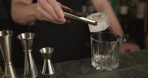 Young male bartender puts an ice cube in a cocktail glass. Close up, pull back.