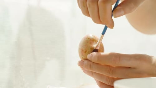 Frau malt zart ein pastellfarbenes Osterei und fügt mit einem Pinsel feine Details hinzu.