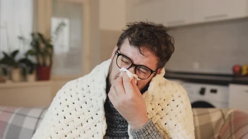 Flu Season Scene Young Man with Glasses Covered in a Duvet Wiping His Nose Seated on Sofa at Home
