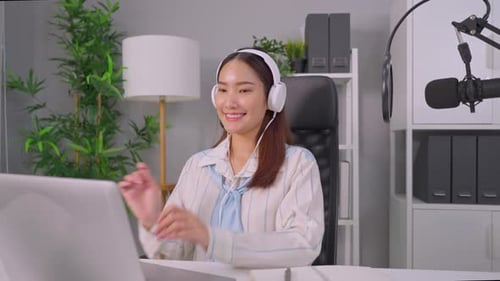 Asian podcaster in headphones using microphone broadcasting in studio.
