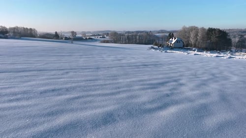 A vast snowcovered field stretches towards a distant house surrounded by trees under a clear blue