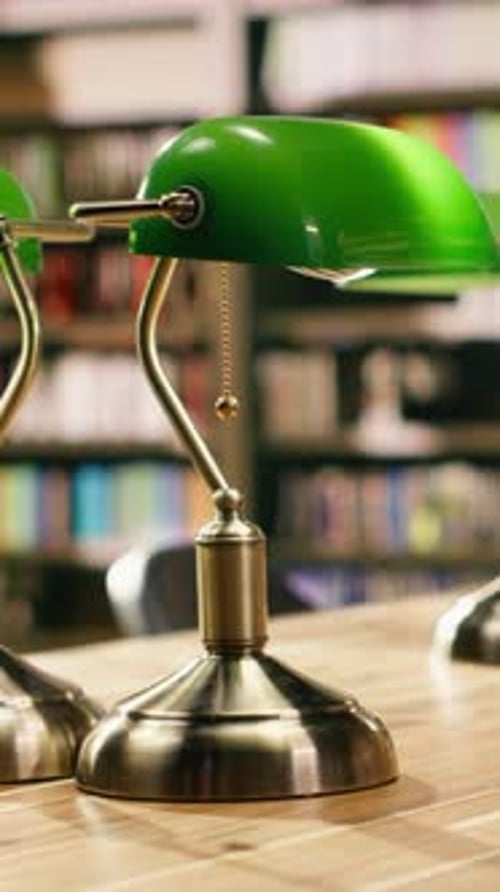 Vertical Video Stack of Books Placed on Wooden Library Desk Beneath Green Lamps