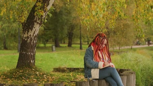 Young Creative Woman with Dreadlocks Drawing Writing in Notebook Sitting on Stumps Near Lake in