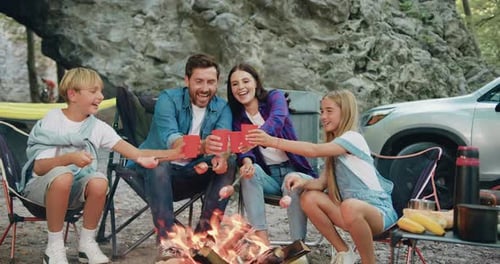 Attaractive family mother and father sitting in forest near rock, roasting sausages by bonfire