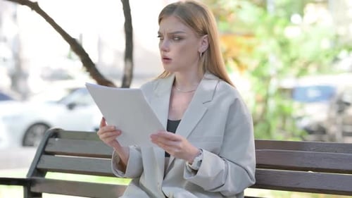 Worried Woman Reviews Paperwork Sitting on Bench