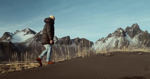 Man Walking On Black Sand Beach With Snowy Mountains