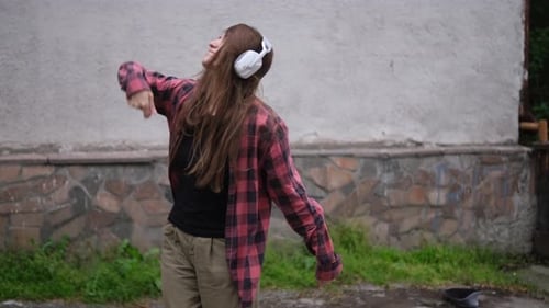 Young Woman in Red Plaid Shirt and Beige Pants Dances Hiphop While Listening to Music Through Large