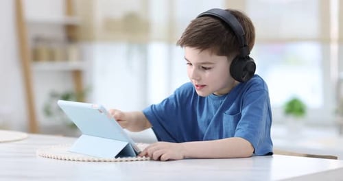 Little boy with headphones using modern tablet at white marble table indoors